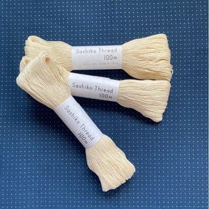 Puede incluir: Tres bobinas de hilo Sashiko color crema, cada una etiquetada "100m", están dispuestas sobre una tela azul oscuro con un estampado de lunares blancos. El hilo está bien enrollado, listo para proyectos de bordado o costura.