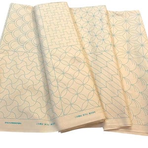 May include: Folded cream-coloured fabric panels featuring light blue stitched geometric patterns. Designs include curved lines, circles, and starburst shapes. The fabric displays detailed stitching and texture, showcasing the intricate patterns.