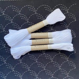 Puede incluir: Cuatro madejas de hilo de bordar blanco agrupadas con etiquetas de papel color canela. Cada etiqueta presenta el texto "SASHIKO" impreso en blanco. El hilo está dispuesto en una pila, mostrando el color blanco brillante y la textura.