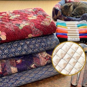 Könnte beinhalten: Ein Stapel aus vier gefalteten Steppstoffen mit roten Blumenmustern, marineblauen geometrischen Formen, violetten Mustern und blauen Wellen. Daneben liegen bunte Stickgarne, ein Nadelkissen und ein runder Ausschnitt mit Rautensteppung.