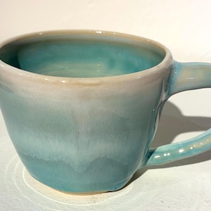 May include: A handmade ceramic mug with a unique, asymmetrical design. The mug features a gradient of colors, transitioning from a light beige rim to a turquoise blue body. The handle is curved and integrated into the mug's design. A stylish addition to any kitchen.