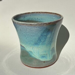 May include: A ceramic tumbler featuring a flared rim and a tapered body. The exterior is finished with a reactive glaze in shades of turquoise, teal, and deep blue, with a visible brown clay base at the bottom edge and rim.
