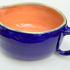 May include: A ceramic bowl with a deep cobalt blue exterior and a smooth, bright orange interior. The bowl features a rounded shape, a single curved handle on the right side, and a small pouring spout on the upper left rim with a light beige edge.