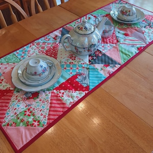 Könnte beinhalten: Ein roter, weißer und blauer Patchwork-Tischläufer mit einer weißen Teekanne, einer weißen Teetasse und einer weißen Untertasse auf einem Holztisch.