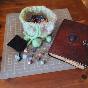May include: A green and pink knitted dice bag with a drawstring closure and pom-poms. The bag is filled with colorful dice and is sitting on a gray cutting mat. There are also several dice scattered on the mat. A brown leather journal with a Celtic design is on the right side of the image.