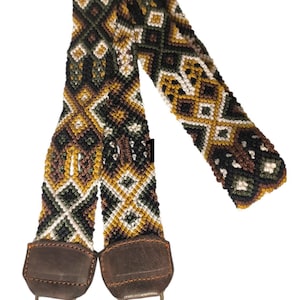 May include: A woven strap with a geometric pattern in shades of brown, green, and white. The strap has two brown leather ends with metal clasps.