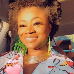 May include: A woman with short, curly, dark brown hair is wearing a white shirt with a colorful abstract print. She is wearing a gold choker necklace and green earrings. The text "Sept. 23" is visible in the image.