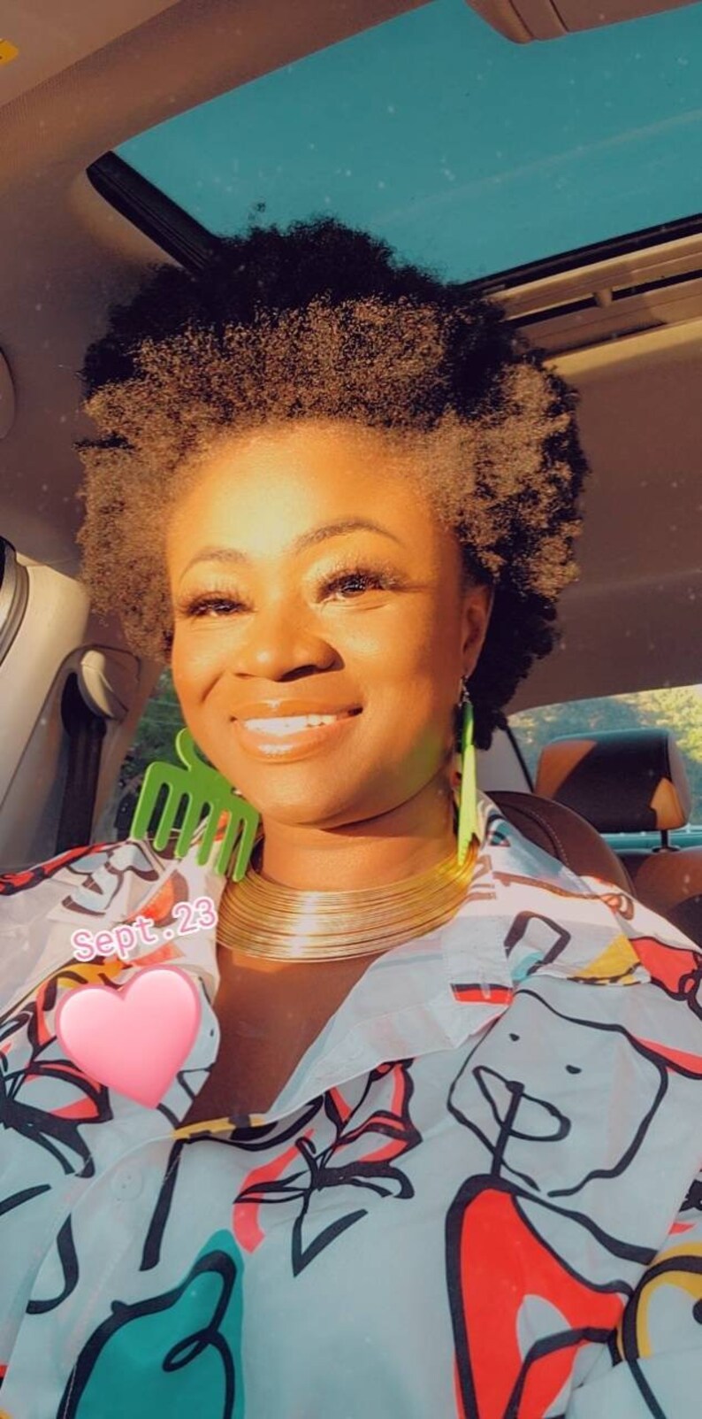 May include: A woman with short, curly, dark brown hair is wearing a white shirt with a colorful abstract print. She is wearing a gold choker necklace and green earrings. The text "Sept. 23" is visible in the image.