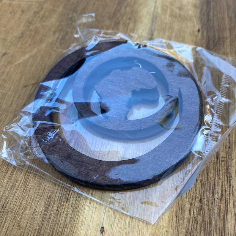 May include: A dark brown wooden ornament shaped like a circle with a cutout of the continent of Africa. The ornament is packaged in a clear plastic bag.