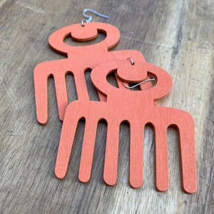 May include: A pair of orange wooden earrings shaped like an Afro comb. The earrings are made of wood and have a smooth finish.