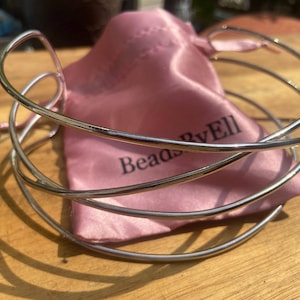 May include: Silver multi-strand bangle bracelet with a pink satin pouch. The pouch has the text "Beads By Ell" printed on it.