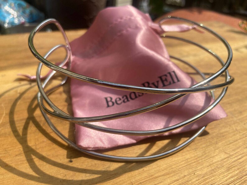 May include: Silver multi-strand bangle bracelet with a pink satin pouch. The pouch has the text "Beads By Ell" printed on it.