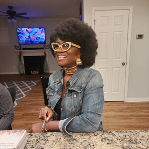 May include: A woman with dark brown hair wearing a blue denim jacket, a black top, and gold jewelry. She is smiling and looking to the left. She is wearing yellow and black cat-eye glasses. The woman is sitting at a table with a granite countertop.