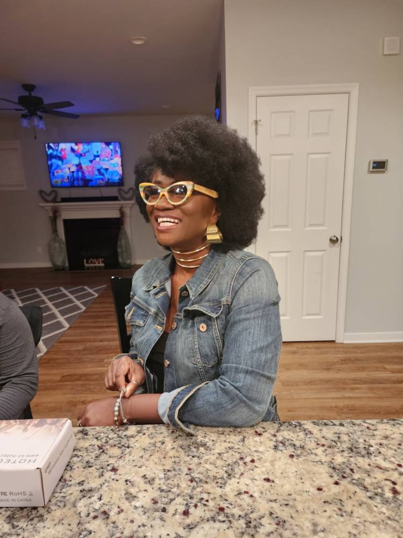 May include: A woman with dark brown hair wearing a blue denim jacket, a black top, and gold jewelry. She is smiling and looking to the left. She is wearing yellow and black cat-eye glasses. The woman is sitting at a table with a granite countertop.