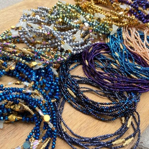 May include: A collection of colourful beaded waist beads, featuring various shades of blue, purple, gold, and silver. The beads are made of glass and have a shiny, iridescent finish. Each strand is adorned with a gold accent.