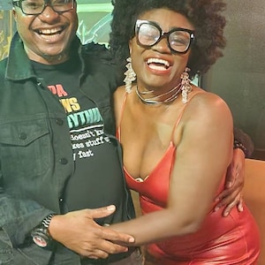May include: Two people are smiling and laughing. The person on the left is wearing a black t-shirt with the words "PA WINS YTHIA" and the words "doesn't by kes stuff u fast". The person on the right is wearing a red leather dress and has large hoop earrings.