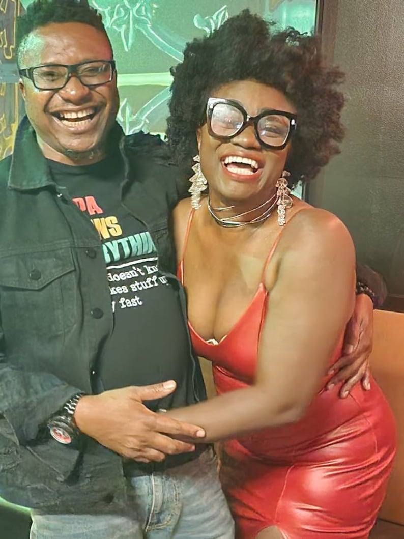May include: Two people are smiling and laughing. The person on the left is wearing a black t-shirt with the words "PA WINS YTHIA" and the words "doesn't by kes stuff u fast". The person on the right is wearing a red leather dress and has large hoop earrings.