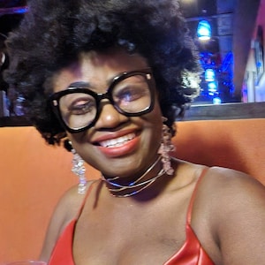 May include: A woman with curly black hair wearing a red sleeveless top, black framed glasses, and a layered gold necklace. She is smiling and looking at the camera.