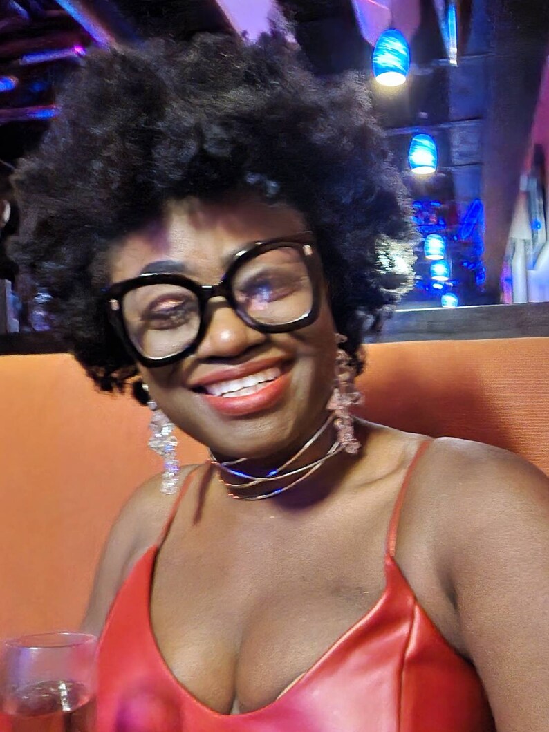 May include: A woman with curly black hair wearing a red sleeveless top, black framed glasses, and a layered gold necklace. She is smiling and looking at the camera.