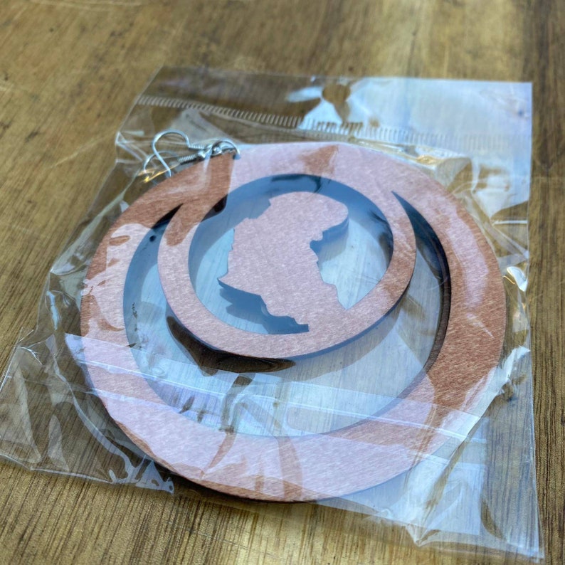 May include: A pair of wooden earrings with a cutout design of the continent of Africa. The earrings are brown and have a circular shape. They are packaged in a clear plastic bag.
