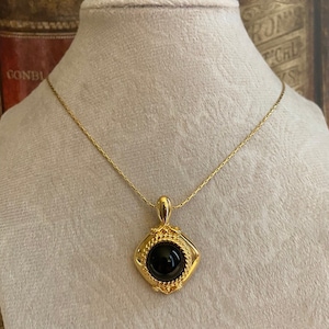 May include: A gold chain necklace with a square pendant featuring a black gemstone. The pendant has a detailed, ornate design.
