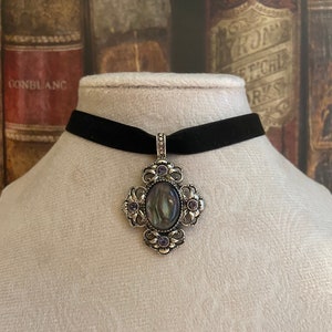 May include: A black velvet choker necklace with a silver pendant featuring an oval iridescent stone surrounded by smaller purple stones.