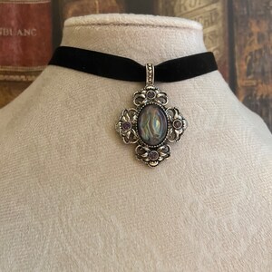 May include: A black velvet choker necklace with a silver pendant featuring a large oval iridescent stone surrounded by smaller purple stones.