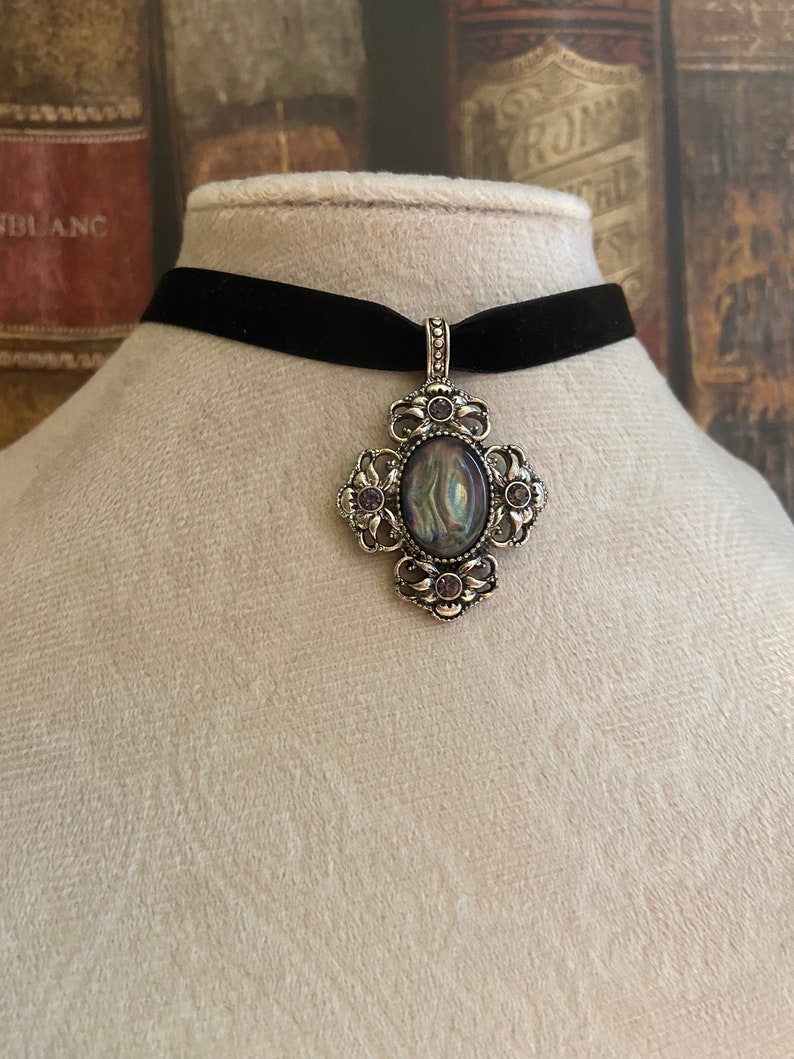 May include: A black velvet choker necklace with a silver pendant featuring a large oval iridescent stone surrounded by smaller purple stones.