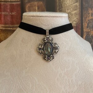 May include: A black velvet choker necklace with a silver pendant featuring an oval abalone shell surrounded by a filigree design and purple rhinestones.