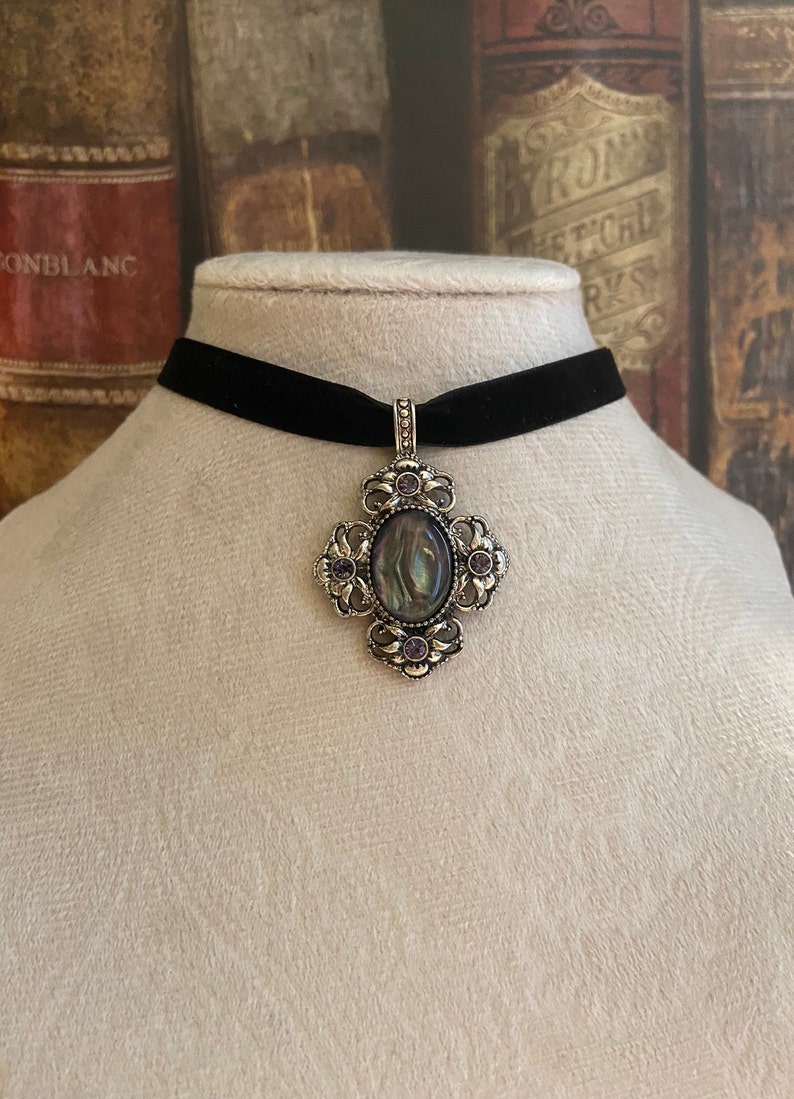 May include: A black velvet choker necklace with a silver pendant featuring an oval abalone shell surrounded by a filigree design and purple rhinestones.