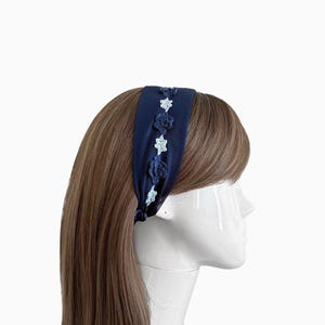 May include: A navy blue headband with a decorative floral design. The headband features a row of crocheted flowers in shades of blue and light blue. The headband is made of a soft, silky material and is designed to be worn around the head.