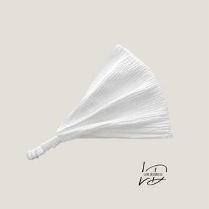 May include: A white headband made of textured muslin fabric featuring a gathered, fan-like design and an elasticized band at the base for a secure fit. The logo for LUXE DESSINS CO is printed in black in the lower right corner of the image.