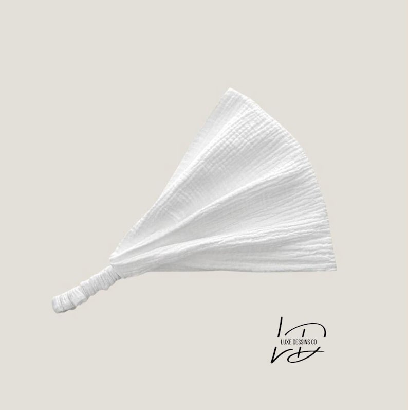 May include: A white headband made of textured muslin fabric featuring a gathered, fan-like design and an elasticized band at the base for a secure fit. The logo for LUXE DESSINS CO is printed in black in the lower right corner of the image.