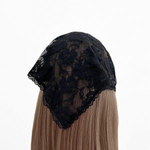 Op de afbeelding: Een zwarte driehoekige hoofddoek van doorschijnende kanten stof met een gedetailleerd bloemenpatroon van rozen. Het accessoire heeft een delicate geschulpte rand langs de omtrek en is gedrapeerd over lichtbruin haar.