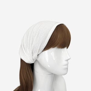 May include: A white head wrap made of a textured, lightweight fabric worn on a mannequin head with brown hair. The fabric features a subtle crinkled pattern and covers the top of the head, extending down to the hairline, providing a simple head covering.