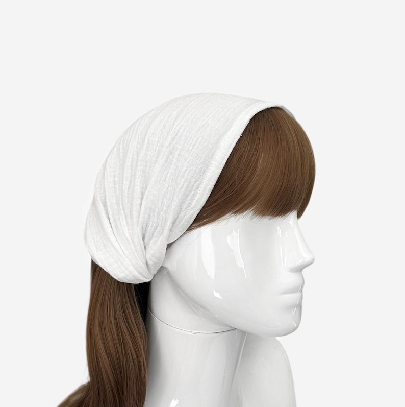 May include: A white head wrap made of a textured, lightweight fabric worn on a mannequin head with brown hair. The fabric features a subtle crinkled pattern and covers the top of the head, extending down to the hairline, providing a simple head covering.