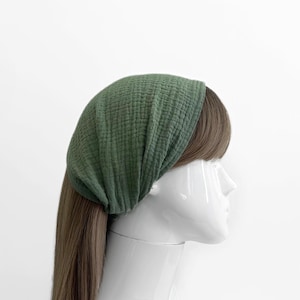 May include: A green headband made of textured fabric. The headband is wrapped around a mannequin head with brown hair. The fabric has a crinkled appearance, and the color is a muted, earthy green.