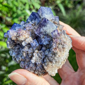 K&ouml;nnte beinhalten: Ein Cluster aus violetten Fluoritkristallen mit einer rauen, nat&uuml;rlichen Textur. Die Kristalle sind in einer dichten Formation zusammengef&uuml;gt, wobei einige kleinere Kristalle die gr&ouml;&szlig;eren umgeben.