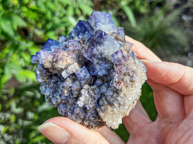 K&ouml;nnte beinhalten: Ein Cluster aus violetten Fluoritkristallen mit einer rauen, nat&uuml;rlichen Textur. Die Kristalle sind in einer dichten Formation zusammengef&uuml;gt, wobei einige kleinere Kristalle die gr&ouml;&szlig;eren umgeben.