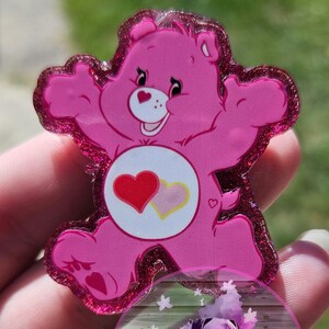 May include: Pink Care Bear with a red and pink heart on its belly. The Care Bear is standing with its arms outstretched. The Care Bear is made of resin and has glitter embedded in it.