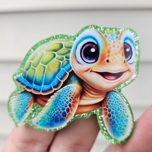 May include: A colorful, cartoon-style sea turtle with a smiling face and large, expressive eyes. The turtle has a green glitter border and a shell with blue, yellow, and green patterns. Its flippers are blue with a dotted pattern.