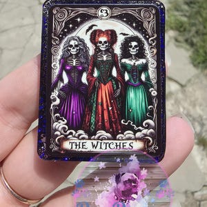 May include: A rectangular, dark-edged card featuring three skeleton witches in colorful gowns. The card reads "THE WITCHES" at the bottom and has a number "3" at the top. The card is held in a hand, with a "BooBerry Designs" logo visible.