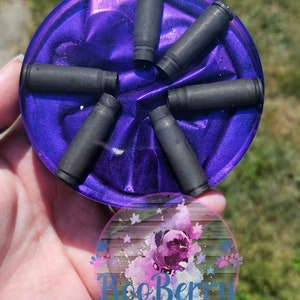 May include: A purple resin coaster with six black bullet casings embedded in the resin. The coaster is round and has a shiny, iridescent finish.