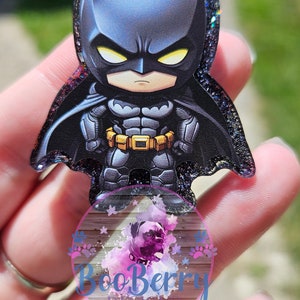 May include: A black and gray glitter resin charm featuring a cartoon depiction of Batman. The charm is shaped like a superhero with a cape and a yellow belt. The charm is held in a hand.