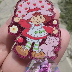 May include: A pink glitter resin badge featuring a cartoon character with a strawberry hat and dress, and a pink cat. The badge has a pink glitter border. The text "BooBerry Designs" is visible on the badge.