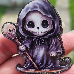 May include: A small, cartoon-style skeleton wearing a purple hooded robe and holding a skull-topped staff. The skeleton has large, round eyes and a wide smile. The image has a glittery background.
