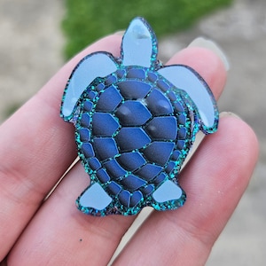 May include: A blue and green glitter resin turtle charm with a white outline. The turtle has a detailed shell pattern.