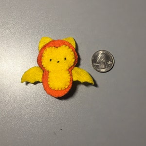 May include: A small, yellow felt bat with orange accents. The bat has a simple, cartoon-like face with black eyes and a stitched smile.