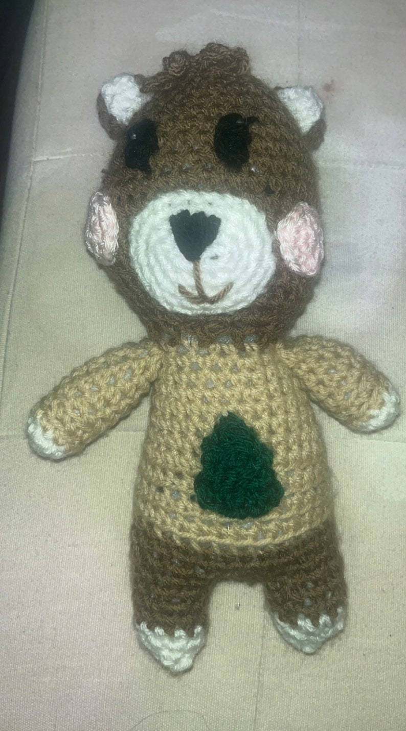 May include: Crocheted bear doll with brown, tan, white, black, and pink yarn. The bear has a white face, black eyes, and a green tree design on its tan sweater. The bear's ears and cheeks are pink.