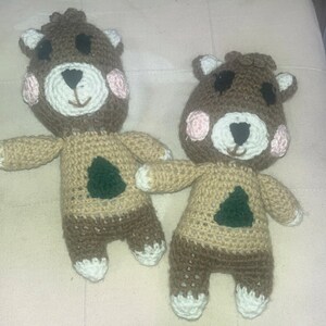 May include: Two handmade crochet bear dolls. The bears have brown heads with white and pink accents, black eyes, and a white muzzle. Their bodies are tan with a brown lower half and a green tree applique. The bears have white paws.